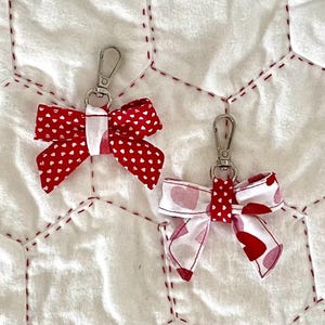 May include: Two fabric bow keychains with silver-toned metal lobster clasps. The left bow is red with a white heart pattern. The right bow is white with a red heart pattern and a red center band featuring small white hearts.