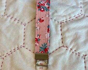 May include: A pink floral key chain with a silver key ring. The fabric has a pink background with red and blue flowers.