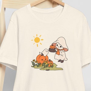 Op de afbeelding: Crèmekleurig T-shirt met een Halloween-illustratie. Het ontwerp toont een cartoongeest, pompoenen en een zon. Een witte baseballcap rust op een houten stoel achter het shirt.