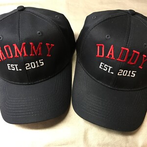 May include: Two black baseball caps with red embroidered text. One cap says "Mommy Est. 2015" and the other says "Daddy Est. 2015".