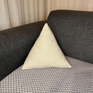 Puede incluir: Un cojín triangular blanco roto con una superficie texturizada descansa sobre un cojín gris y blanco estampado. El cojín está colocado sobre un sofá gris oscuro, con una cortina de color neutro en el fondo. La forma del cojín añade un toque moderno.