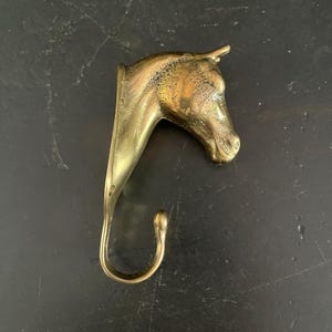 May include: A brass wall hook shaped like a horse head. The metallic object features a detailed equine profile with a curved hook extending from the base of the neck. The surface has a polished gold-toned finish with natural patina and dark accents.