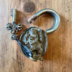 Peut inclure: Un cadenas métallique avec une anse ouverte et une clé assortie. La face avant du cadenas présente un relief sculpté représentant une divinité chevauchant un animal. La clé possède une poignée ornée et complexe. Un cordon noir relie la clé au cadenas.