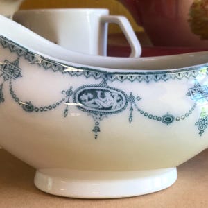 May include: White porcelain gravy boat with a curved spout and a loop handle. The boat is decorated with teal blue floral and geometric patterns. The design includes a central oval medallion with a figure. A classic piece for serving sauces.