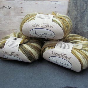 May include: Three skeins of Lighthouse Hayward Pointe yarn in shades of beige, brown, and olive green. Each skein has a paper label with product information. The yarn is arranged on a dark gray surface.