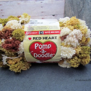 May include: A skein of Red Heart Pomp-a-Doodle yarn in a mix of colors including cream, brown, and olive green. The yarn is super bulky and has a pom-pom texture. The label reads "Pomp-a-Doodle" and "Super Bulky".