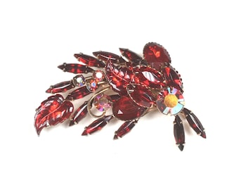 Pode incluir: Um broche de strass vermelho em forma de flor estilizada com uma pedra central iridescente.
