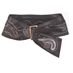 May include: A wide black belt featuring a textured surface with silver-toned snake-like patterns. The belt includes a smooth black leather-like trim along the top edge and a rectangular metallic buckle in a muted gold finish at the centre.