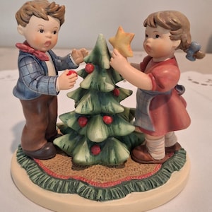 May include: A ceramic figurine featuring a young child in a blue plaid jacket and brown pants decorating a green Christmas tree with red ornaments, while another child in a red dress places a yellow star on top, all set on a round base.
