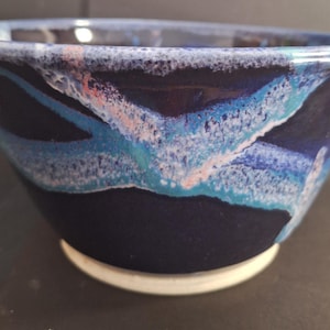 May include: A ceramic bowl with a dark blue exterior and a white base. The interior and rim feature a blend of blue, teal, and pink hues. The bowl's design has a unique, artistic appearance, suitable for serving food or as a decorative piece.