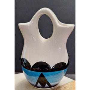 May include: A white ceramic vase with a handle, featuring a design of black, blue, and white geometric shapes. The vase has a rounded base and a wide opening. The design includes a blue band and black mountain-like shapes.