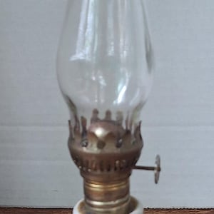 May include: A vintage oil lamp with a clear glass chimney and a ceramic base. The base is white with brown and blue floral designs. The lamp has a brass-colored metal fitting and a small adjustment knob.