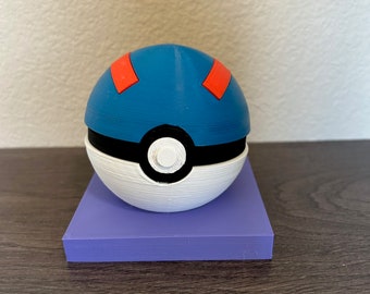 May include: A 3D-printed Poké Ball replica, featuring a white bottom half, a black band, and a blue top with two orange rectangular accents. The ball sits on a solid purple rectangular base, with a textured surface.