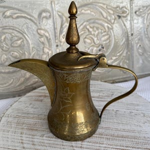 Puede incluir: Una tetera de latón con un diseño floral adornado. La tetera tiene un pico, una manija y una tapa con un pomo.