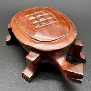 Könnte beinhalten: Eine braune, schildkrötenförmige Holzschatulle mit glatter, polierter Oberfläche. Der Panzer weist in der Mitte ein erhabenes, gitterartiges Quadratmuster auf. Das Objekt hat vier geschnitzte Beine und einen Kopf, der aus dem Körper herausragt und die natürliche Holzmaserung zeigt.