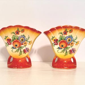 May include: Two matching ceramic vases with a fan-shaped top and a flared base. The vases are painted with a floral design in shades of orange, yellow, blue, and red. The base is a deep orange color, and the top has a scalloped edge.