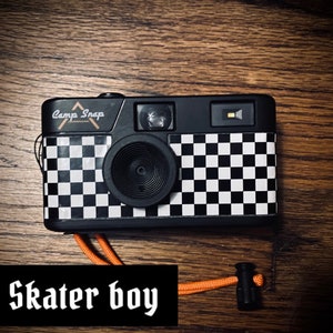 May include: A black and white checkered camera with a black strap and the words "Camp Snap" printed on the front. The camera is sitting on a wooden surface.