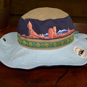 Peut inclure: Un chapeau de soleil beige et bleu clair orné d'un paysage désertique peint à la main avec des formations rocheuses rouges et des montagnes enneigées. Il comporte une bande décorative à motif géométrique et une étiquette Hand Painted by Sheri Wiseman Art.