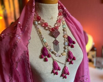 May include: A mannequin head displays layered necklaces with pink and clear beads, and fuchsia teardrop pendants. A sheer pink scarf with sequin trim drapes around the neck. The jewelry is set against a neutral background.
