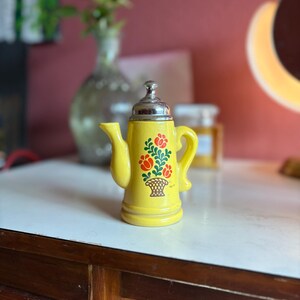 May include: A yellow ceramic teapot with a silver lid and a floral design. The teapot has a red, green, and orange floral pattern with a basket in the center.