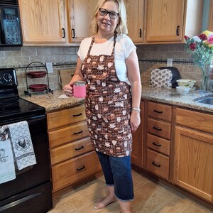 May include: A woman wearing a brown apron with a coffee cup pattern, holding a pink mug. The apron features various coffee cup designs on a brown background. She is standing in a kitchen with wooden cabinets and a black stove.