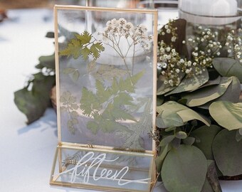 May include: A gold framed glass table number with pressed greenery and white flowers inside. The table number is set on a wooden slice surrounded by eucalyptus greenery.