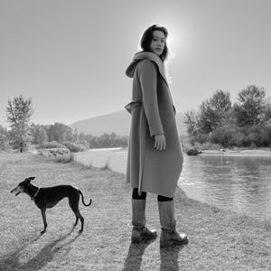 Puede incluir: Una mujer con un abrigo marrón con capucha y botas marrones se encuentra junto a un río con un perro negro. La mujer mira por encima del hombro a la cámara.