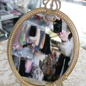 May include: A gold-colored oval mirror with a pearl-like trim. The mirror has a decorative top with a jeweled design and the word "Allah" in Arabic script. The mirror is sitting on a white lace surface.
