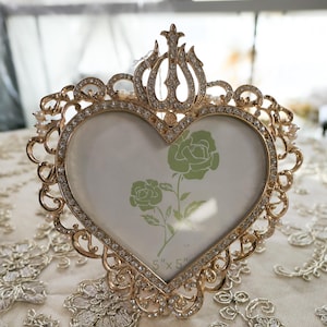May include: Gold heart-shaped picture frame with a decorative design and rhinestones. The frame is 5 inches by 5 inches and has a clear glass insert.