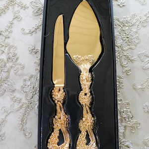 May include: A gold-colored cake knife and server set in a black box. The handles are ornate with a floral design.