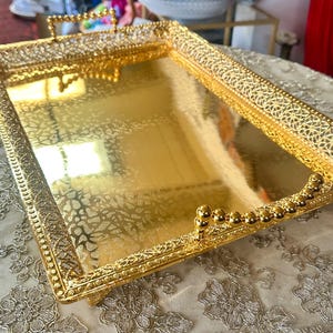 May include: A rectangular gold-colored tray with ornate filigree detailing around the edges and handles. The tray has a reflective surface with a subtle pattern. The handles are adorned with small, gold-colored spheres.