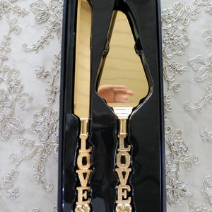 May include: Gold wedding cake knife and server set in a black box. The handles are engraved with the word "LOVE" and have a heart at the bottom.