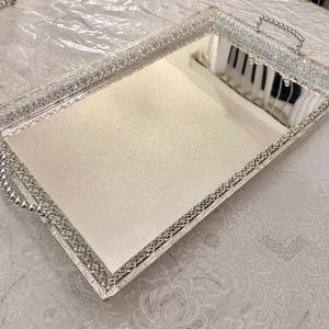 May include: A rectangular silver mirrored tray with ornate filigree detailing around the edges and handles. The tray reflects the surrounding environment, creating a shimmering effect. The handles are adorned with small, round embellishments.