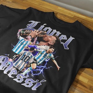 May include: Black t-shirt featuring a graphic of a soccer player with the name "Lionel Messi" in a stylized font. The design includes images of the player in action, with lightning bolt accents.
