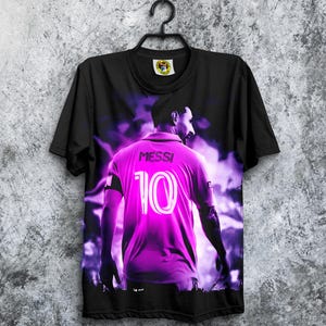 May include: Black t-shirt featuring a vibrant image of a soccer player in a purple jersey with the name "MESSI" and the number "10" on the back. The design has a purple and white color scheme, with a dynamic, artistic background.