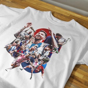 May include: White t-shirt featuring a collage of soccer players, including a central image of a smiling player. The design incorporates a red circle and a blue circle, with various action shots of players in different uniforms.