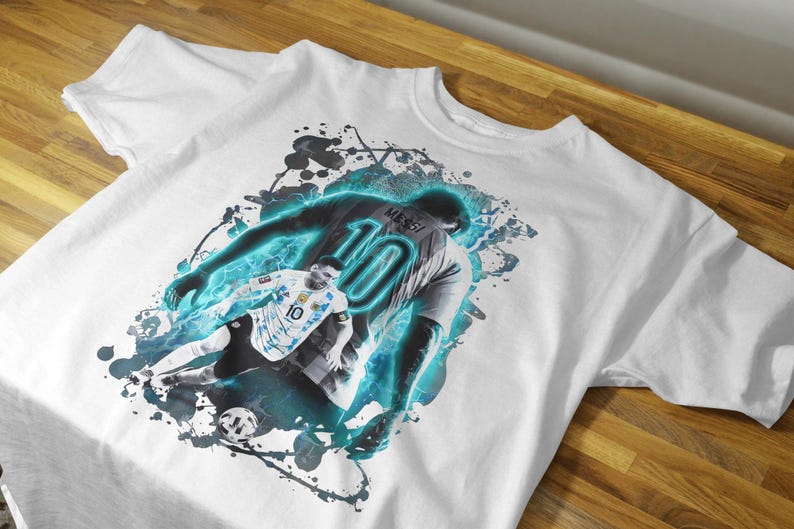 May include: White t-shirt featuring a graphic of a soccer player in action, with the number 10 on the jersey. The design includes blue and black accents, and the player's name is visible. The shirt is displayed on a wooden surface.