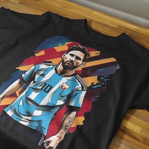 May include: Black t-shirt featuring a stylized illustration of a soccer player in a light blue and white striped jersey with the number 10. The graphic includes a colorful background and a portrait of the player.