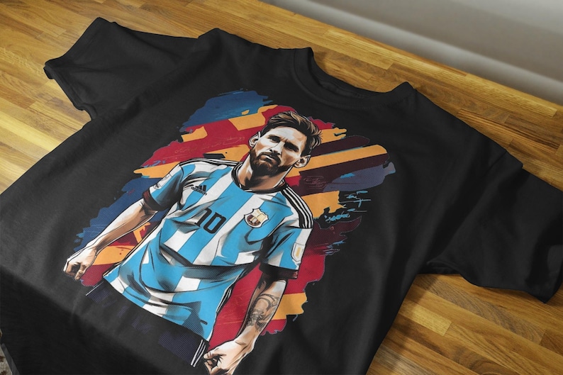 May include: Black t-shirt featuring a stylized illustration of a soccer player in a light blue and white striped jersey with the number 10. The graphic includes a colorful background and a portrait of the player.