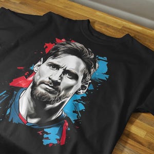 May include: Black t-shirt featuring a stylized portrait of a man with a beard. The portrait is rendered in shades of gray, with splashes of red and blue. The shirt is displayed on a wooden surface.