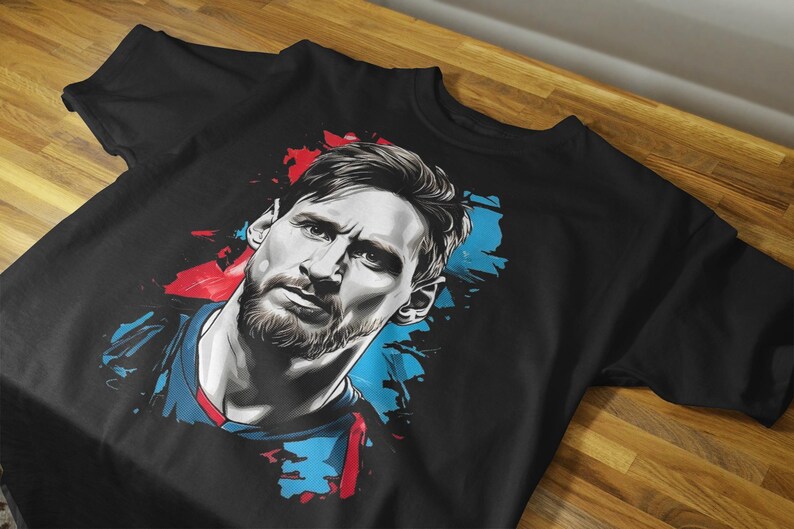 May include: Black t-shirt featuring a stylized portrait of a man with a beard. The portrait is rendered in shades of gray, with splashes of red and blue. The shirt is displayed on a wooden surface.