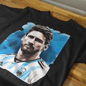May include: Black t-shirt featuring a portrait of a man with a beard, set against a blue and white background. The man is wearing a white and light blue striped jersey with a crest. The shirt is displayed on a wooden surface.