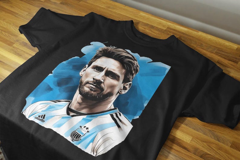 May include: Black t-shirt featuring a portrait of a man with a beard, set against a blue and white background. The man is wearing a white and light blue striped jersey with a crest. The shirt is displayed on a wooden surface.