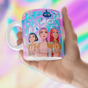 May include: White ceramic mug with the text "K-POP DEMON HUNTERS" and illustrations of three anime-style women. The mug has a white handle and a colourful, swirling background with a cat graphic.