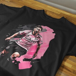 May include: Black t-shirt featuring a stylized illustration of a soccer player in a pink and white uniform. The graphic has a pink and black watercolor effect. The word "butleii" is visible on the shirt.