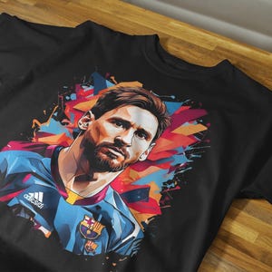 May include: Black t-shirt featuring a colorful graphic of a soccer player. The design includes a portrait of the player with a blue and red color scheme. The shirt has the Adidas logo.