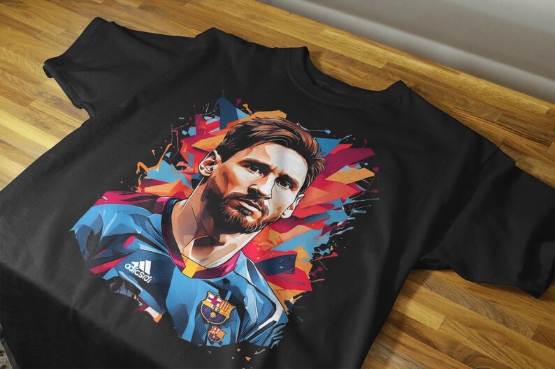 May include: Black t-shirt featuring a colorful graphic of a soccer player. The design includes a portrait of the player with a blue and red color scheme. The shirt has the Adidas logo.