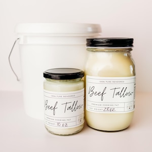 Op de afbeelding: Twee glazen potten met 100% puur gerenderd rundvet, een pot van 10 oz en een pot van 28 oz. Beide potten hebben zwarte deksels en witte etiketten met de tekst "100% puur gerenderd rundvet" en "Premium kookvet".