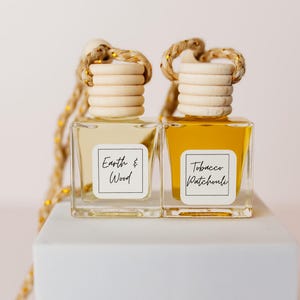 以下が含まれることがあります： 木製の栓と金色の紐がついた小さなガラス瓶が2本。瓶は透明な液体と琥珀色の液体で満たされています。瓶には「Earth & Wood」と「Tobacco Patchouli」と書かれたラベルが付いています。