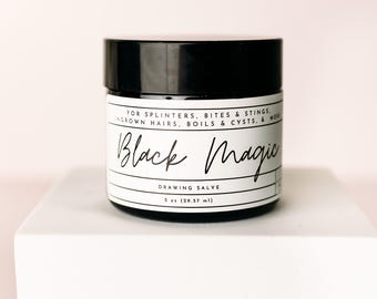 Op de afbeelding: Een pot Black Magic zalf met een zwart deksel en een wit etiket. Het etiket bevat de tekst "FOR SPLINTERS, BITES & STINGS, INGROWN HAIRS, BOILS & CYSTS, & MORE" en "DRAWING SALVE". De pot bevat 29,57 ml product.